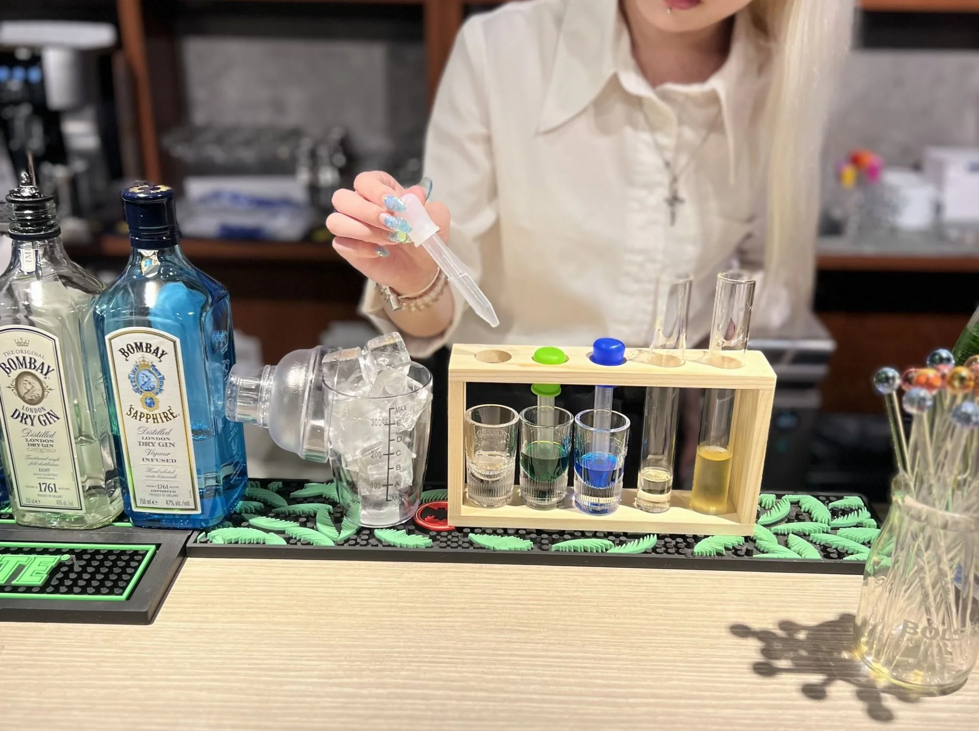 白いシャツのスタッフがスポイトで色のついたドリンクを調合している、試験管立てとシェイカーが並ぶカウンター